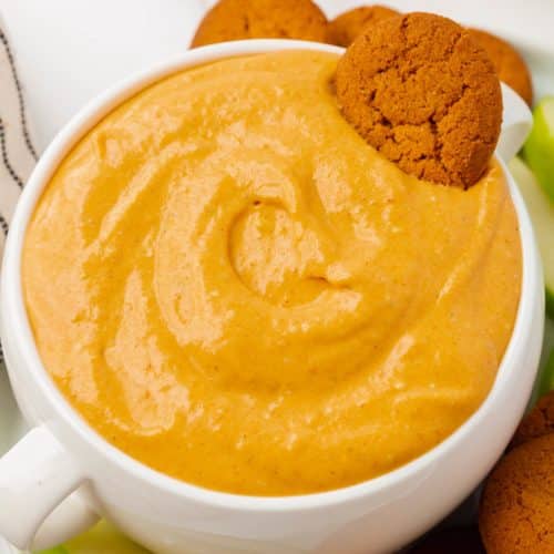 A large bowl with two handles, filled with creamy pumpkin dip. A gingersnap is in the bowl of dip. Under the bowl is a white plate filled with more gingersnaps and sliced green apples.