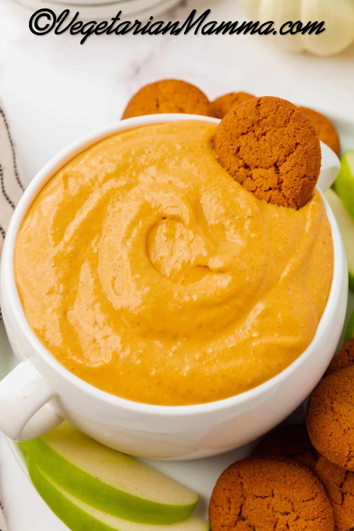 A large bowl with two handles, filled with creamy pumpkin dip. A gingersnap is in the bowl of dip. Under the bowl is a white plate filled with more gingersnaps and sliced green apples.