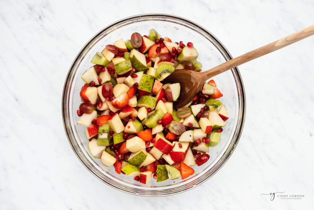 a glass bowl filled with thanksgiving fruit salad. A wooden spoon is stirring it together.