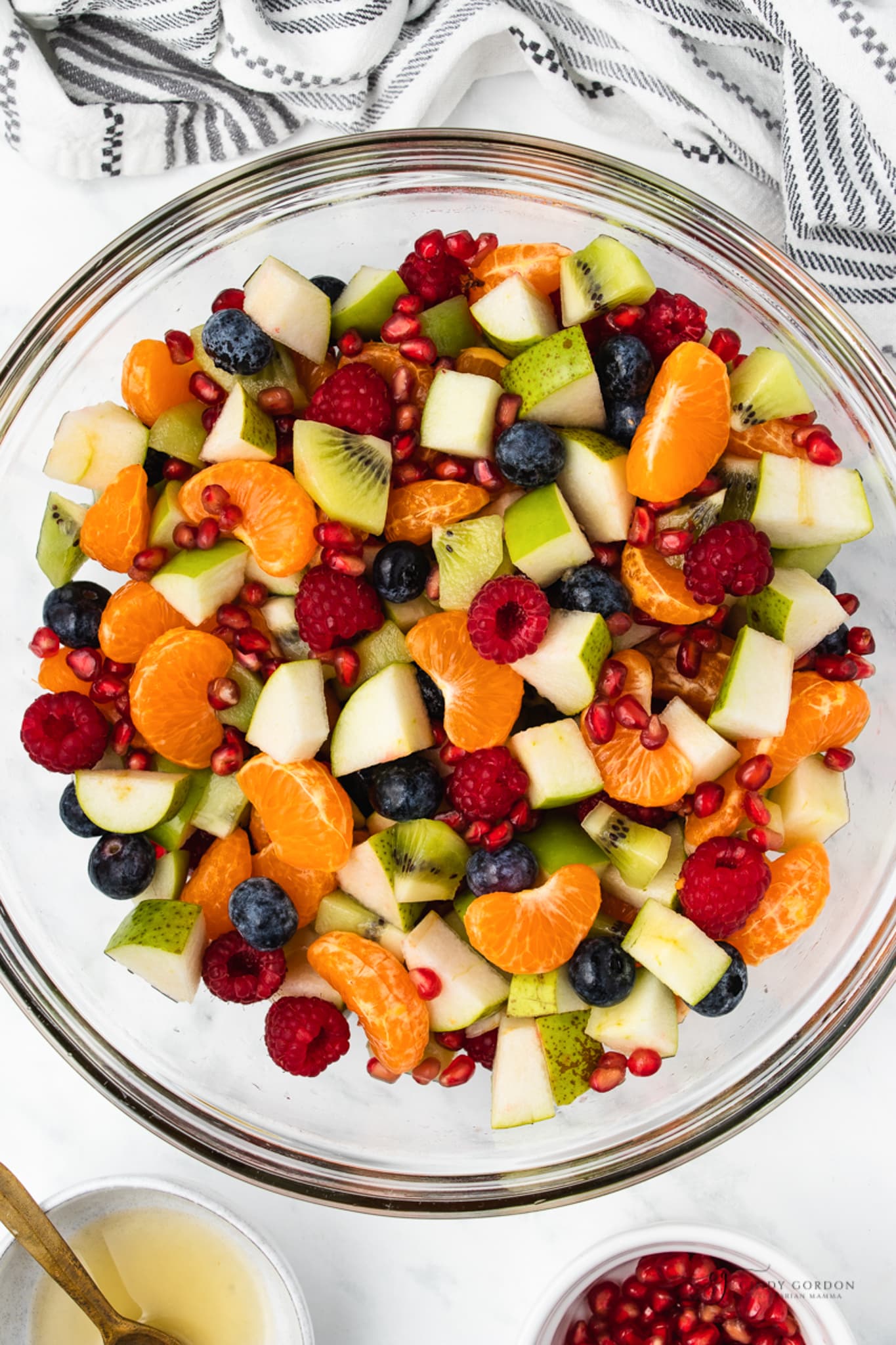 a clear glass bowl filled with fruit salad featuring clementines, green apples, and pomegranate seeds
