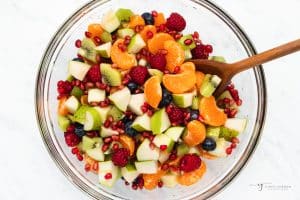 a clear glass bowl filled with fruit salad featuring clementines, green apples, and pomegranate seeds. A spoon is stirring it all together.