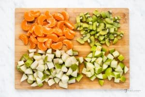 Tangerines sectioned, on a cutting board with chopped apples, chopped pears, and chopped kiwi fruit