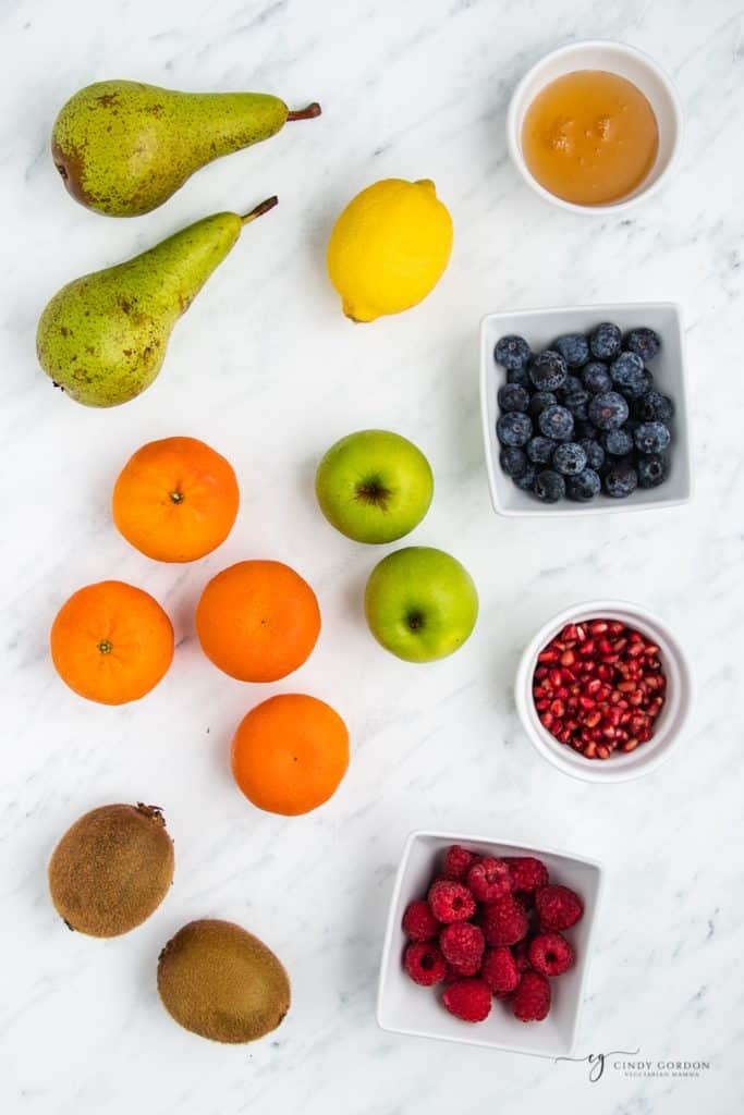 Fruits on a counter, being prepared for fruit salad. Includes green pears, green apples, kiwis, tangerines, blueberries, raspberries, and pomegranate seeds.