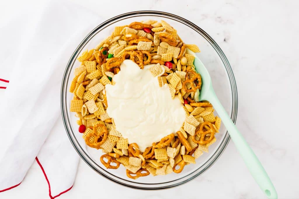melted white chocolate poured into a bowl of chex and pretzels. A spatula is about to stir it in.