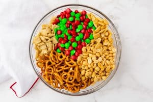holiday M&Ms, peanuts, chex ceral, and pretzels in a clear glass bowl.