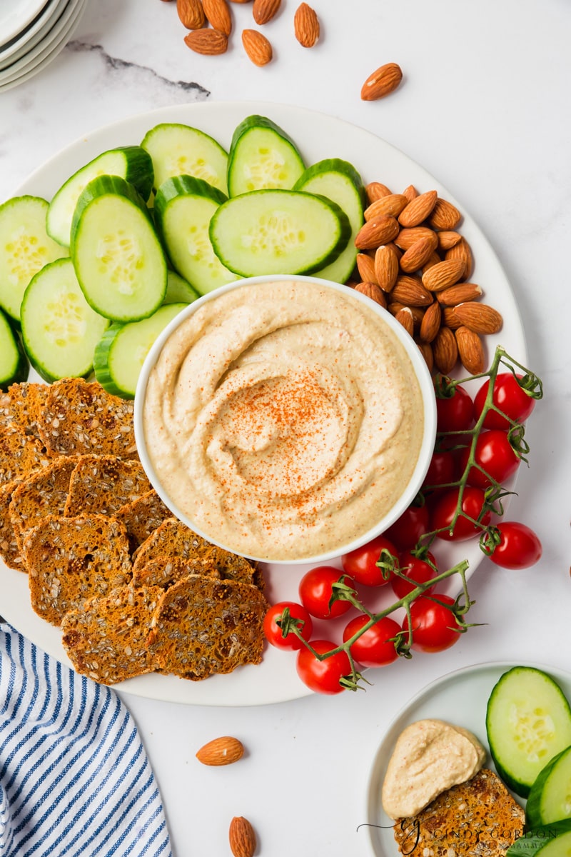top down view of a plate of sliced cucumbers, tiny tomateos, almonds, and seed crackers. In the center of the plate is a bowl of creamy Bitchin sauce. 