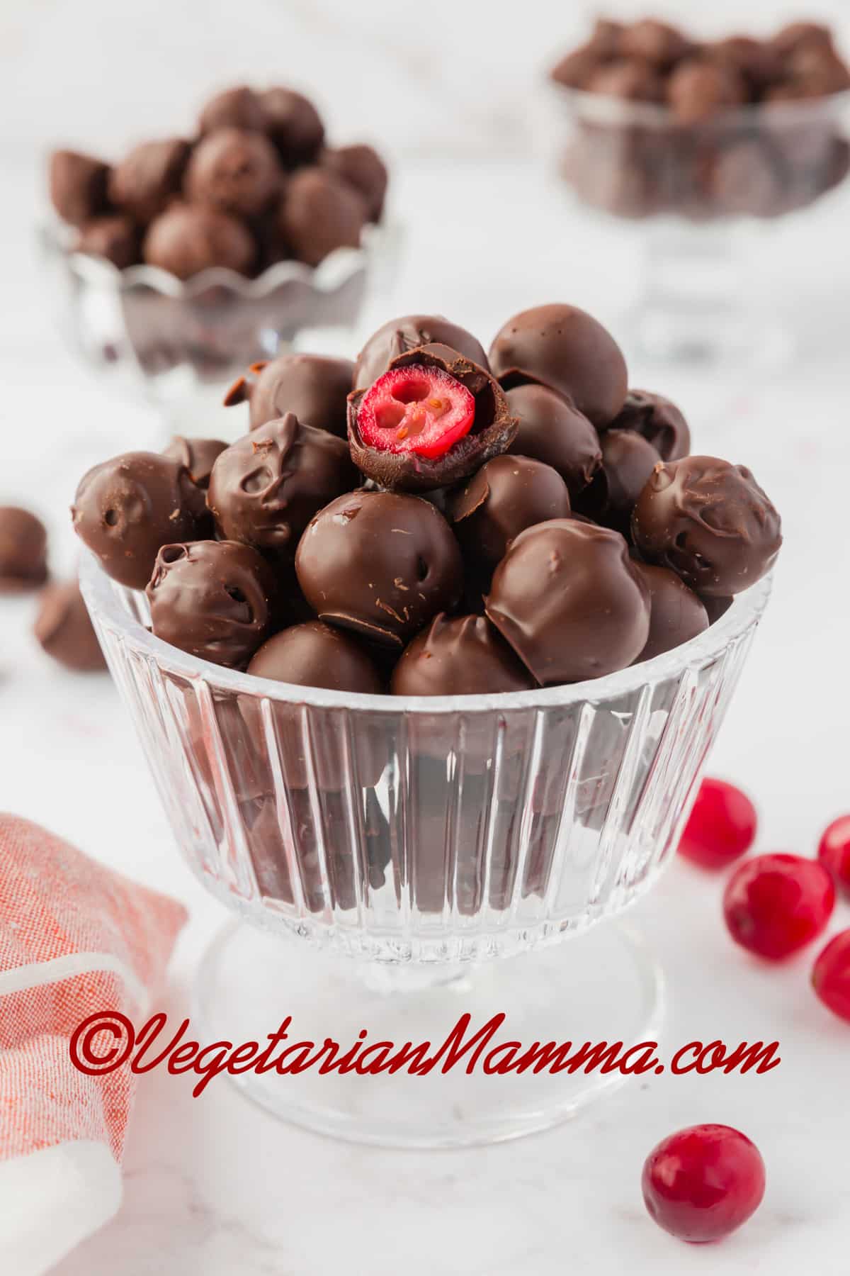 A footed glass dessert dish filled with chocolate-covered cranberries. One has been bitten into to show the inside.