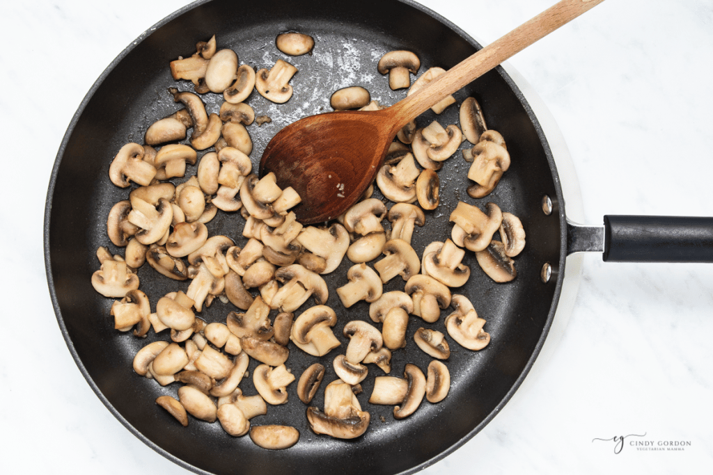 sliced mushrooms sauteed in a frying pan with a wooden spoon.