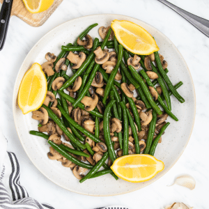 a round white plate filled with green beans and mushrooms. Three lemon wedges are around the edge of the plate.