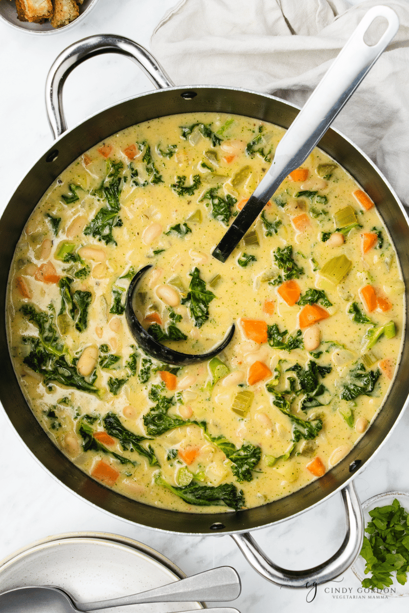 Partially blended white bean and kale soup in a pot. A silver ladle is in the pot, and dishes are around.