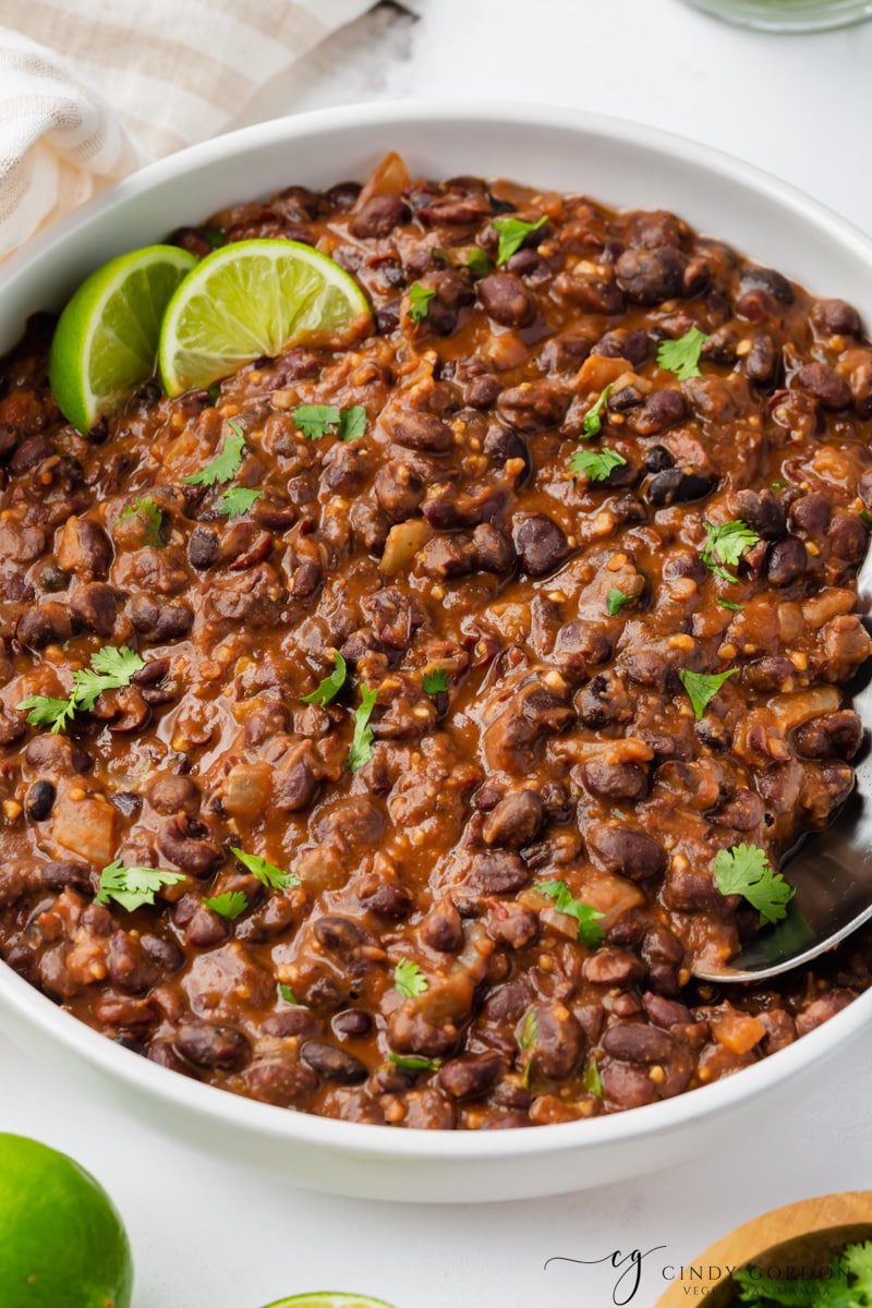 a large white bowl of seasoned black beans garnished with two lime wedges and chopped cilantro. A spoon is in the bowl.