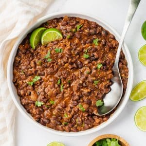 a large white bowl of seasoned black beans garnished with two lime wedges and chopped cilantro. A spoon is in the bowl.