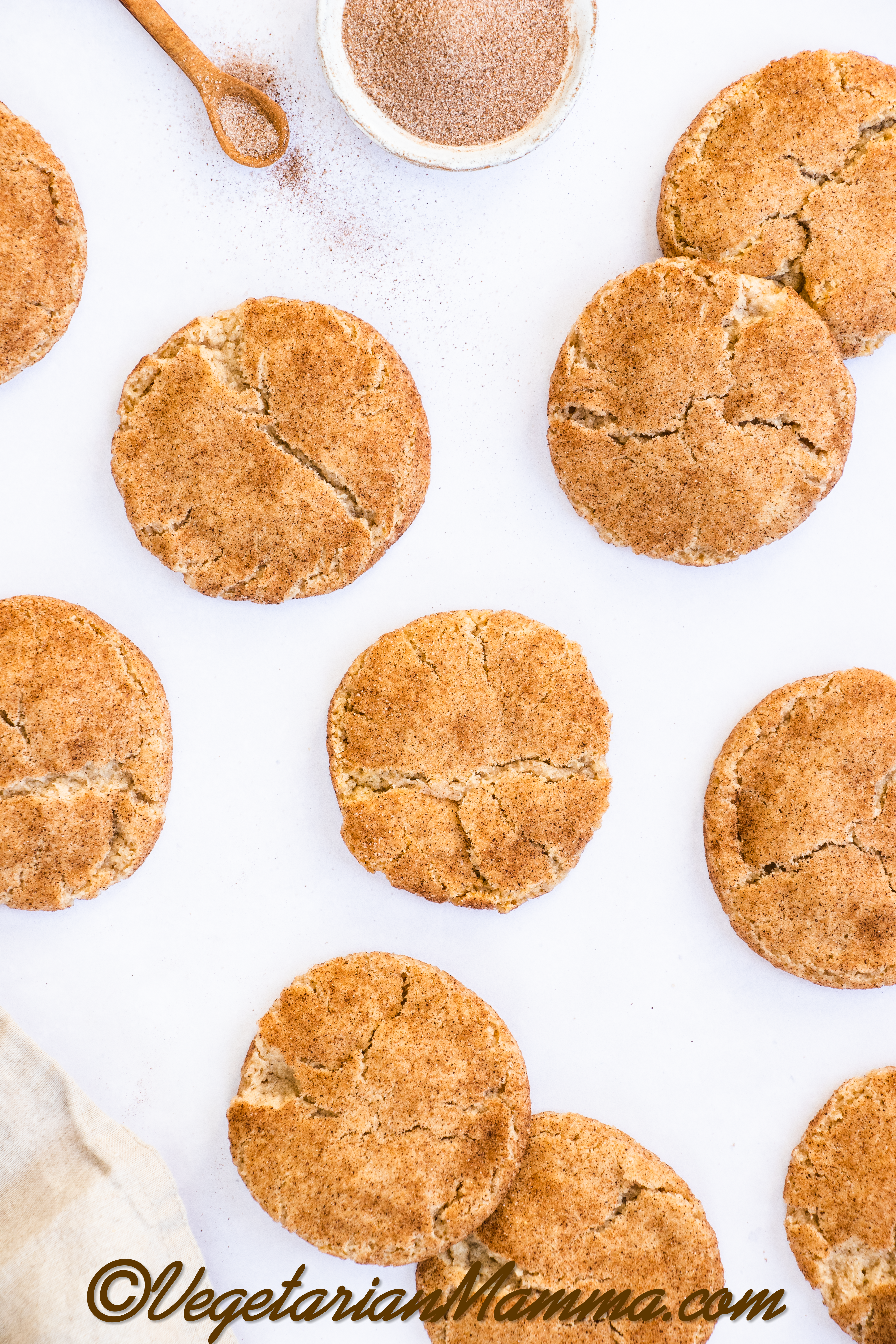 An array of snickerdoodle cookies on a white countertop next to a bowl and a spoonful of cinnamon sugar