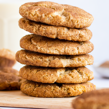 A stack of 6 snickerdoodle cookies on a wooden cutting board surrounded by more cookies