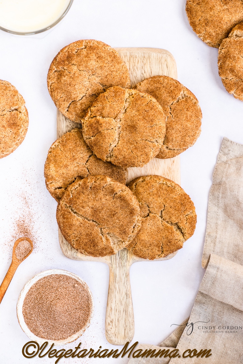 Six large snickerdoodle cookies on a wooden cutting board surrounded by more cookies and a towel