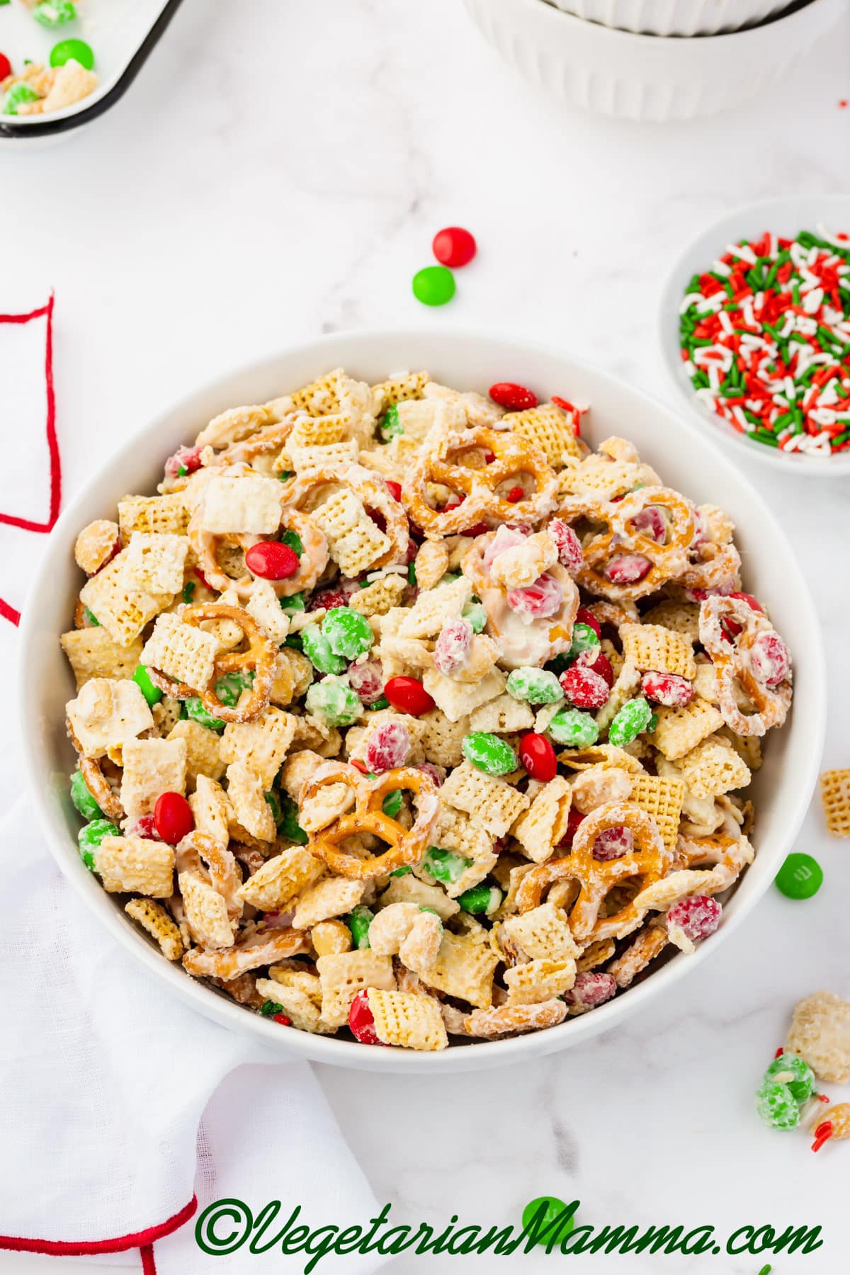 White bowl of red and green M&Ms, melted chocolate, peanuts, pretzels, chex pieces.