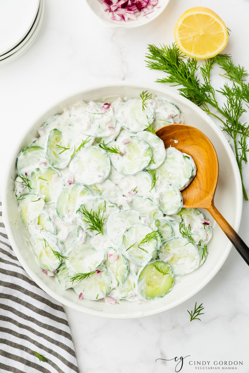 A large white bowl filled with sour cream polish cucumber salad and a wooden spoon. The salad is viewed from directly overhead, and sprigs of fresh dill and a halved lemon are next to it on the counter. 
