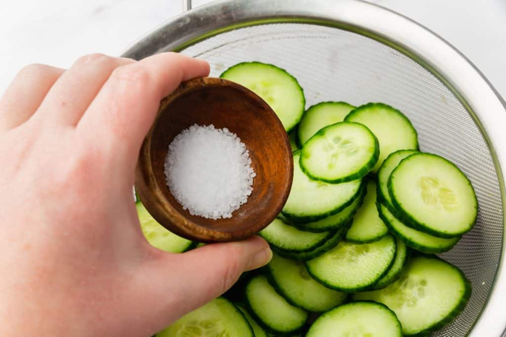a hand pouring a small bowl of salt into a colander full of cucumber slices.