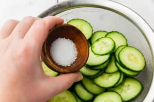 a hand pouring a small bowl of salt into a colander full of cucumber slices.