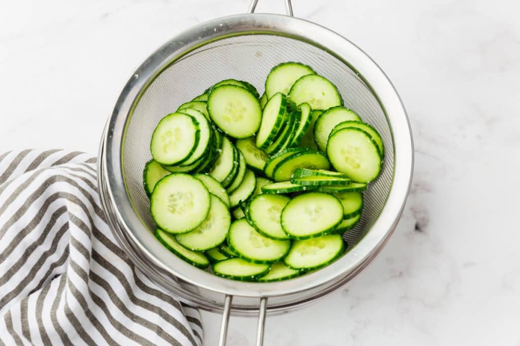 Salted cucumber slices in a mesh sieve, draining.