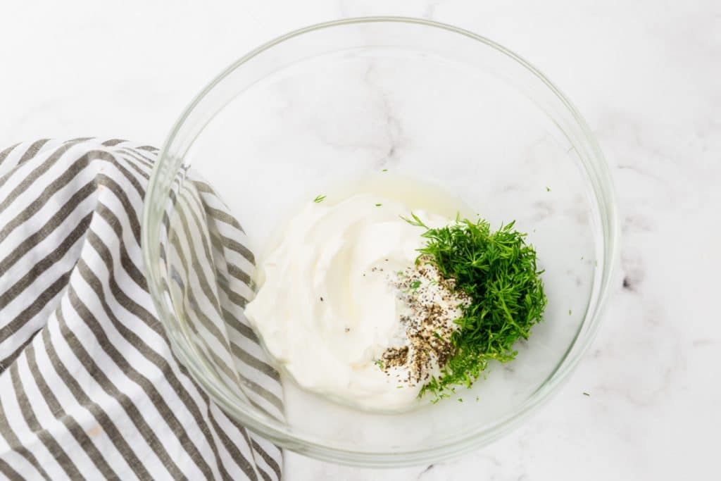 A glass mixing bowl with sour cream and seasonings for cucumber salad.