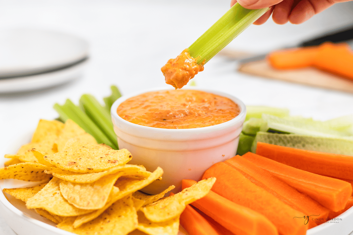 a platter of tortilla chips, carrot sticks and celery sticks with a large cup of orange red pepper dip in the middle. a piece of celery is being dipped in the red pepper crema. 