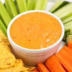 a bowl of orange roasted red pepper crema dip, surrounded by carrot sticks, celery sticks and chips.