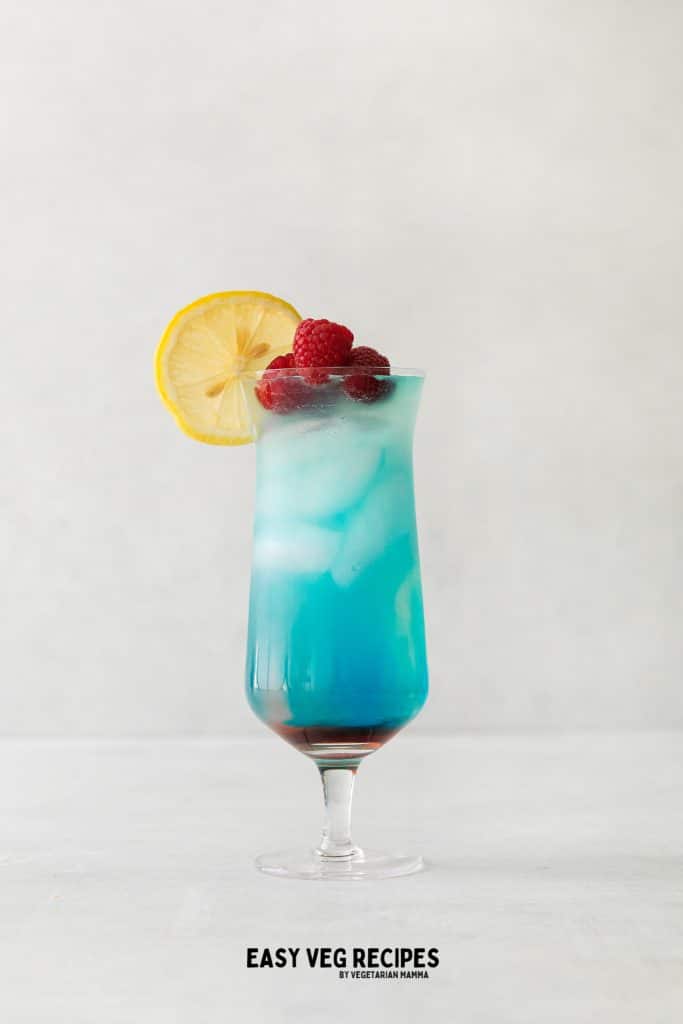 Grateful Dead Drink, in a tall clear glass. Red liquid on bottom, most of the liquid is blue and ice cubes in it. Lemon wheel and raspberries on top