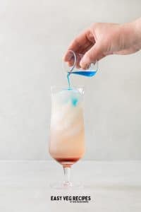 blue liquid being poured into glass with ice cubes and red liquid