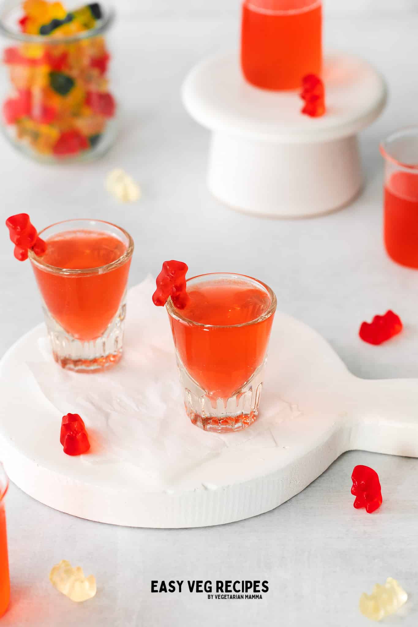 shot glasses filled with red liquid and a red gummy bear on the side. a jar of colorful gummy bears to the back left. Lots of gummy bears on table top