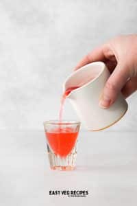 hand pouring a white jar full of red liquid into shot glass