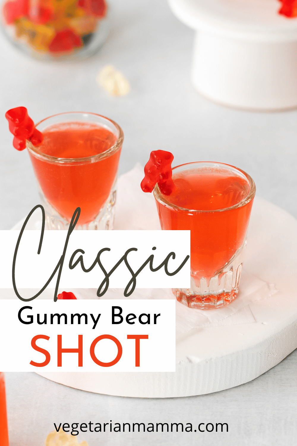 shot glasses filled with red liquid and a red gummy bear on the side. a jar of colorful gummy bears to the back left. Lots of gummy bears on table top text overlay: classic gummy bear shot