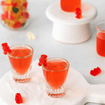 shot glasses filled with red liquid and a red gummy bear on the side. a jar of colorful gummy bears to the back left. Lots of gummy bears on table top