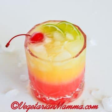a highball glass with a layered red and yellow malibu sunset drink. Garnished with a cherry and lime slices.