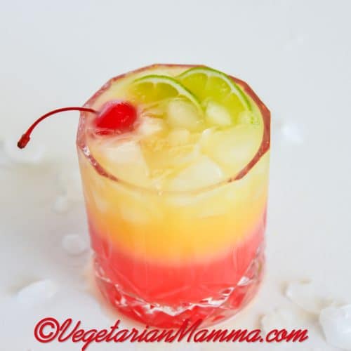 a highball glass with a layered red and yellow malibu sunset drink. Garnished with a cherry and lime slices.