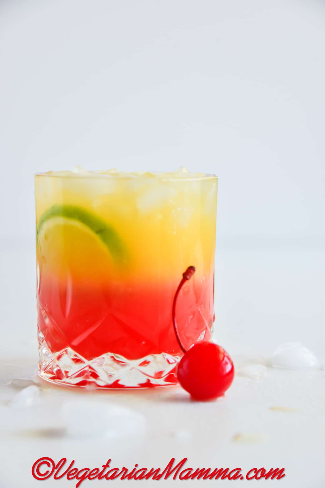 a highball glass with a layered red and yellow malibu sunset drink. Garnished with a cherry and lime slices.