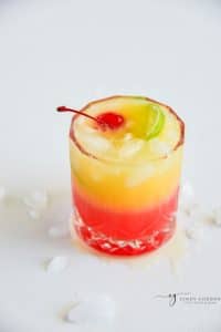 a rocks glass with a layered red and yellow malibu sunset drink. Garnished with a cherry and lime slices.