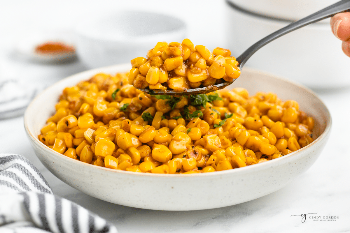 A large white serving bowl filled with cajun fried corn. A spoon is lifting up a serving. 