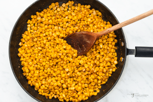 A skillet of yellow corn kernels and cajun seasonings, stirred with a wooden spoon.
