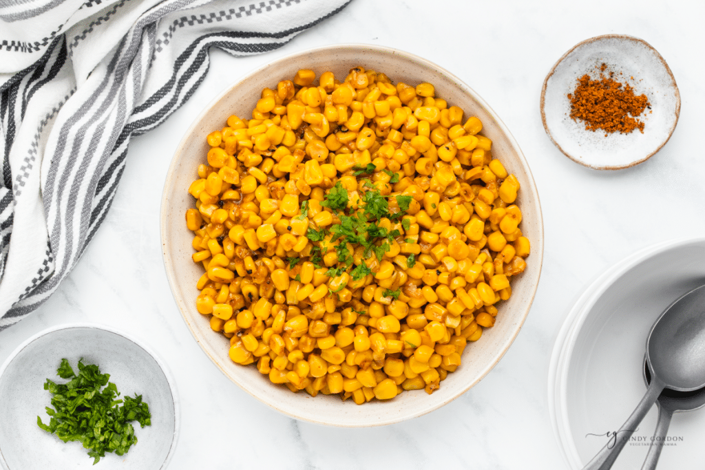 a large ceramic serving bowl filled with yellow corn with cajun seasonings and parsley garnish. There is a spoon under the bowl and a small plate of cajun seasoning above it. View is from overhead.