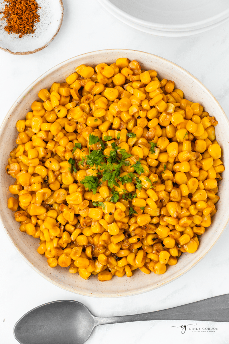 a large ceramic serving bowl filled with yellow corn with cajun seasonings and parsley garnish. There is a spoon under the bowl and a small plate of cajun seasoning above it. View is from overhead. 