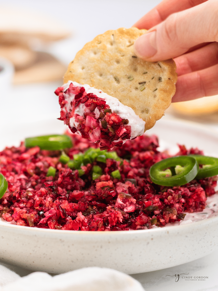 a hand holding a large round cracker and dipping it into chopped cranberry jalapeno dip. 