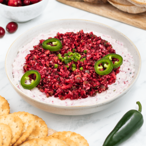 a bowl of cranberry dip with jalapenos on a table next to crackers.