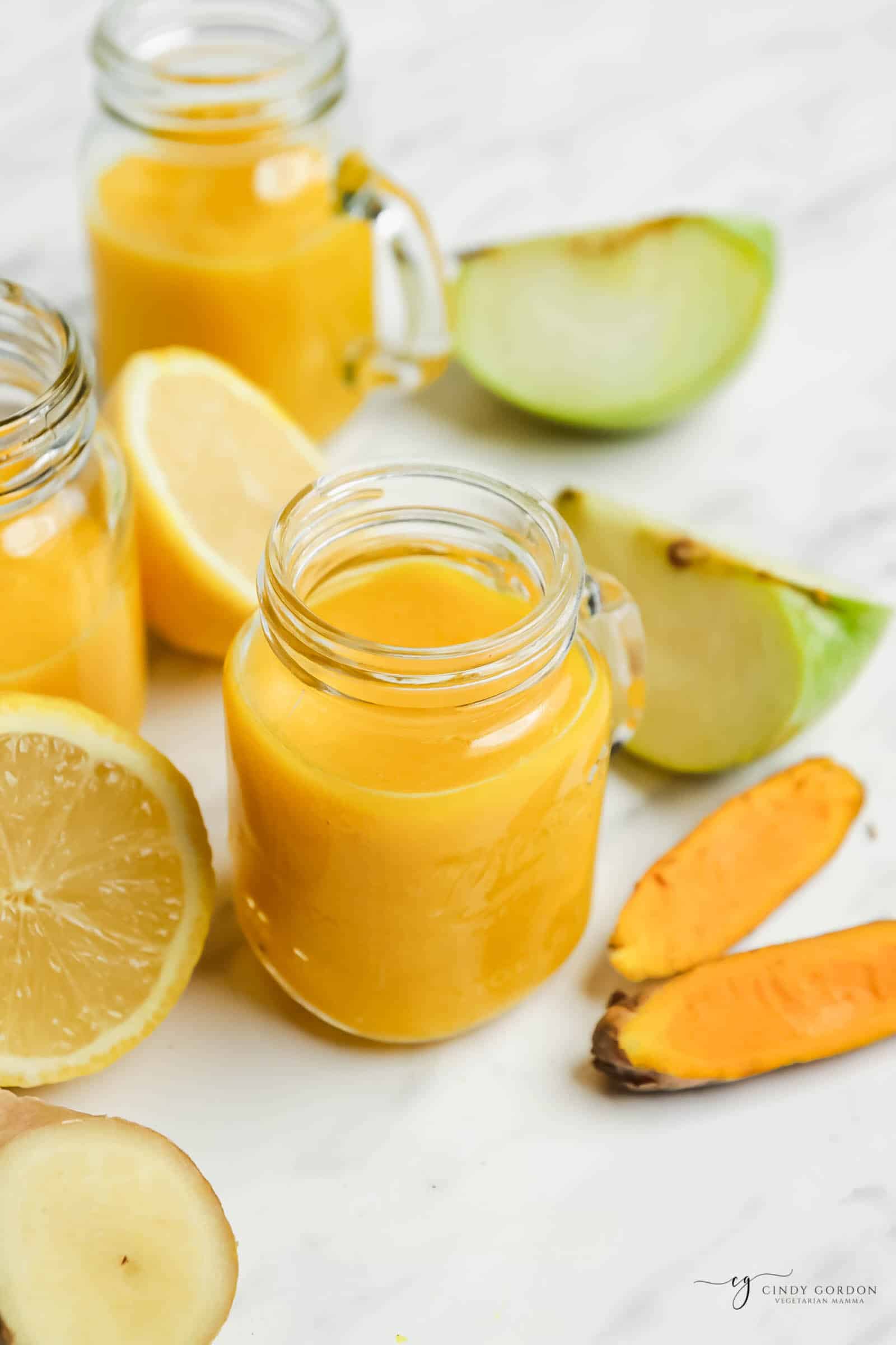 a small mason jar mug filled with orange ginger tumeric juice. Around the jar are pieces of apples, lemon, ginger, and tumeric root