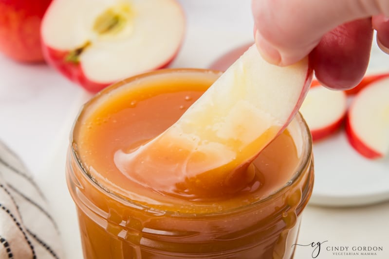 An apple slice dipping into a small glass jar filled with homemade microwave caramel sauce