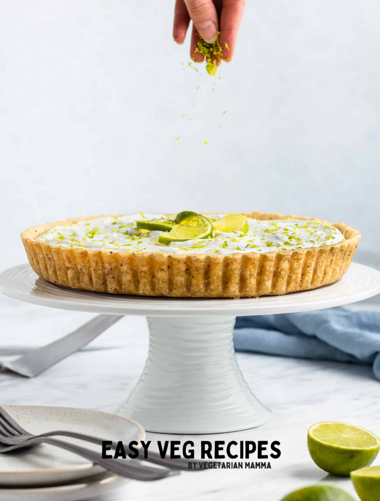 A key lime pie on a white cake pedestal. A hand is sprinkling something over it.
