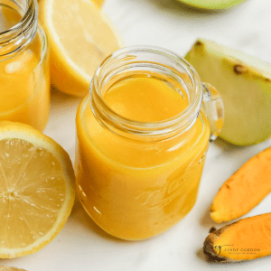 a small mason jar mug filled with orange ginger turmeric juice. Around the jar are pieces of apples, lemon, ginger, and turmeric root