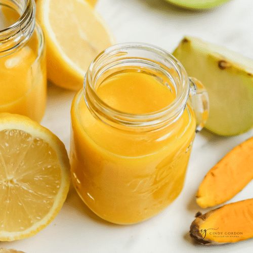 a small mason jar mug filled with orange ginger turmeric juice. Around the jar are pieces of apples, lemon, ginger, and turmeric root