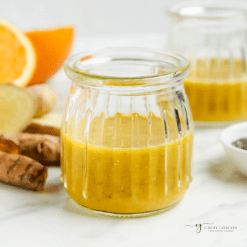 two glass jars holding homemade wellness shots. In the background are oranges, ginger, and turmeric roots