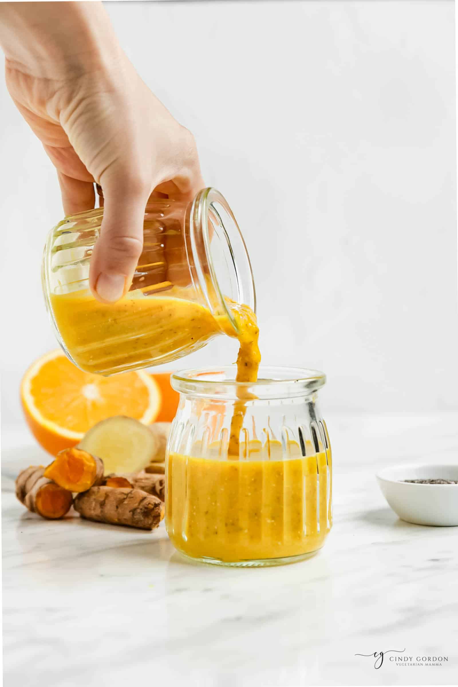 a hand pouring a jar of orange wellness shot into a second jar. In the background are fresh turmeric and ginger.
